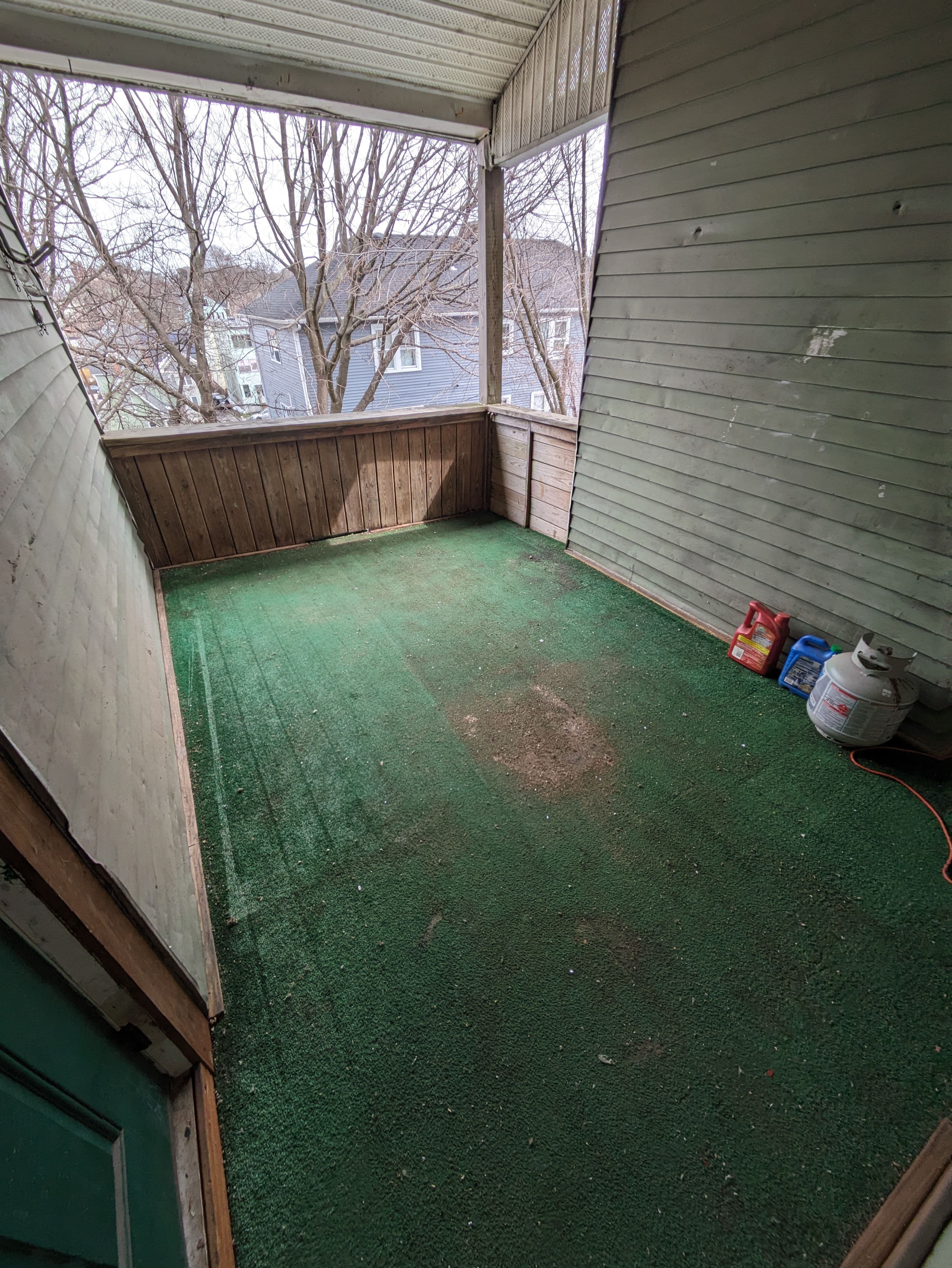 Empty porch with green carpet, wooden walls, and a view of surrounding trees and houses.
