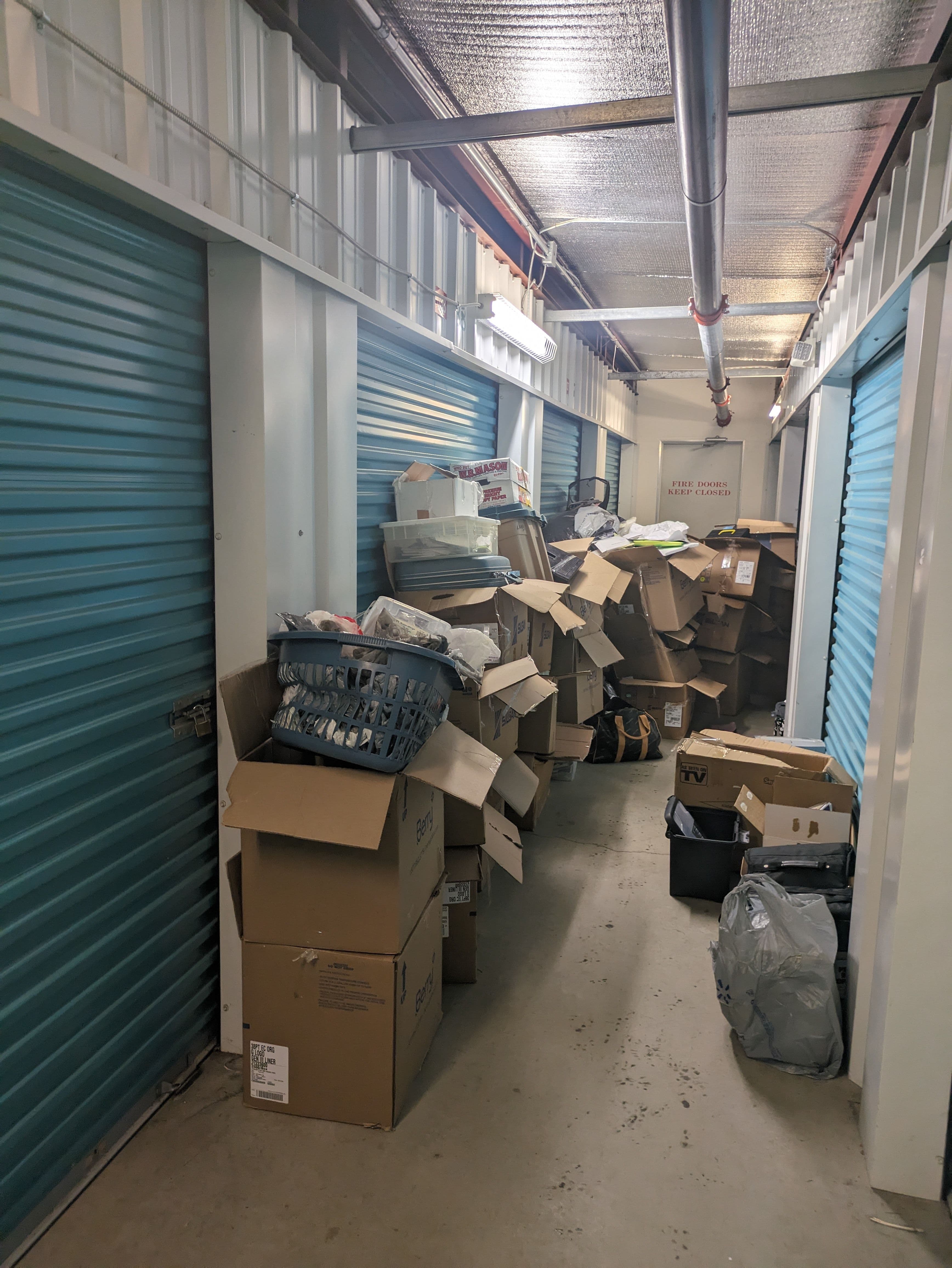 Cluttered storage unit with stacked boxes and items in a narrow hallway.